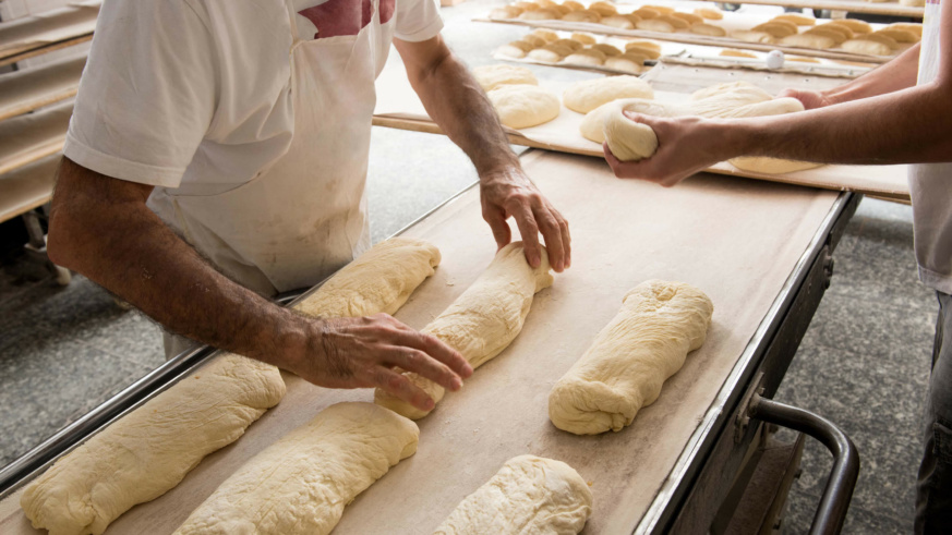 "Bakers shaping dough into loaves on a long table in a bakery setting."