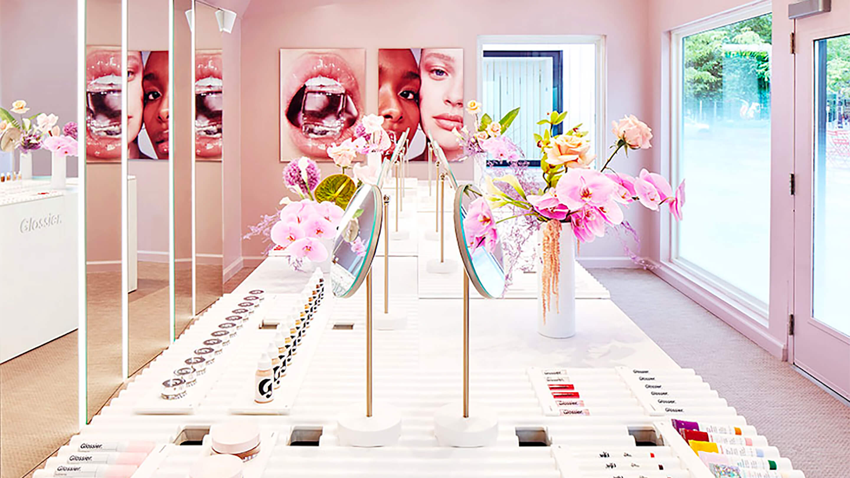 Inside a glossier store with an assortment of flowers, mirrors and makeup placed on a white table