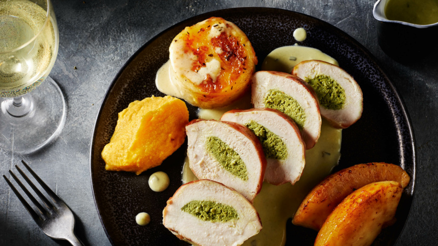 "Sliced chicken breast stuffed with herb filling, accompanied by potatoe fondate and vegetables on a black plate."