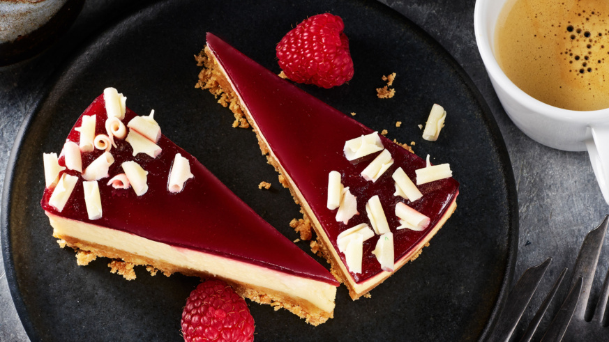 "Two slices of raspberry cheesecake topped with white chocolate curls, garnished with fresh raspberries."